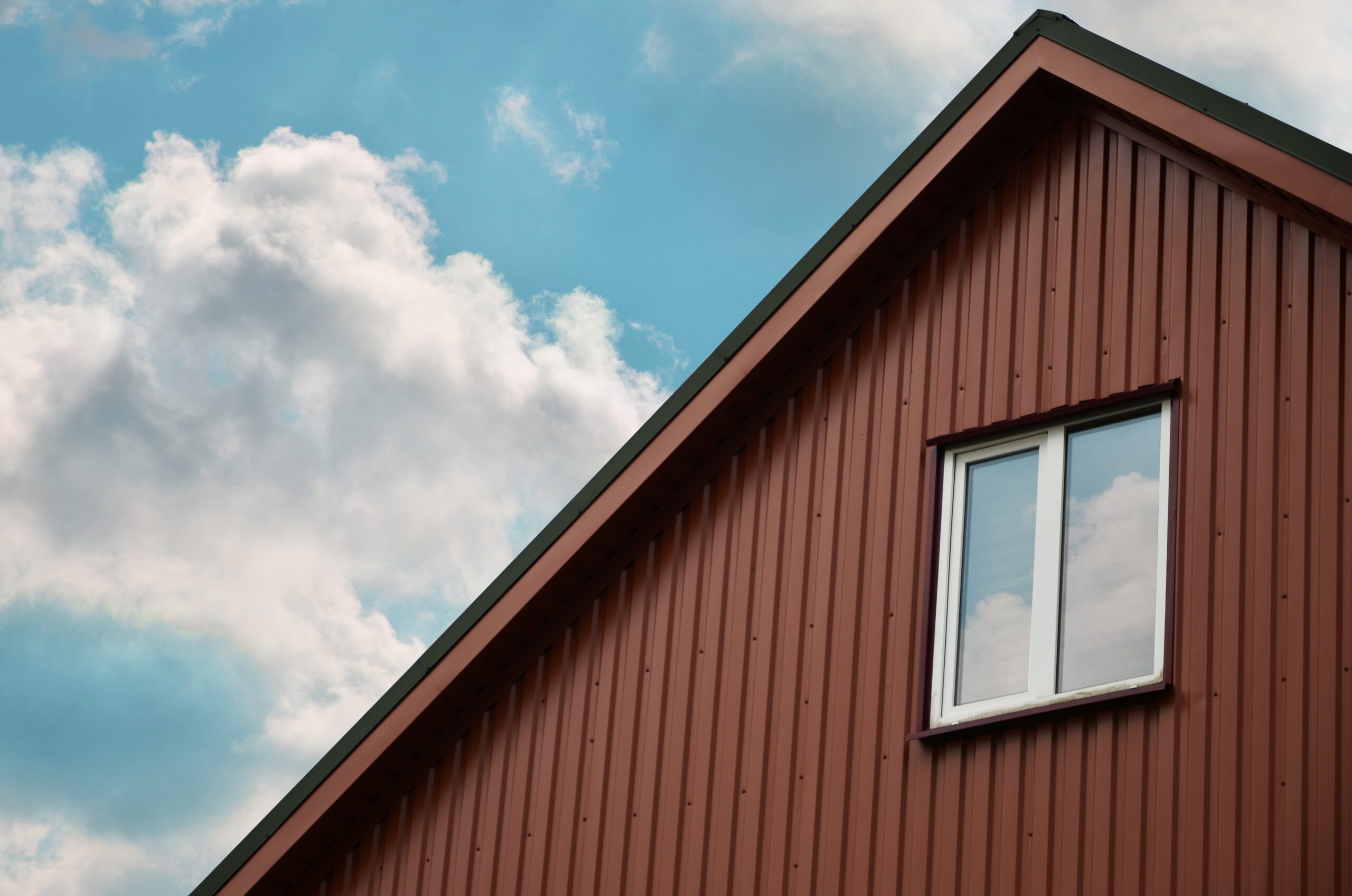 The gable end of a house with brown metal siding and a window, set against a blue sky with clouds