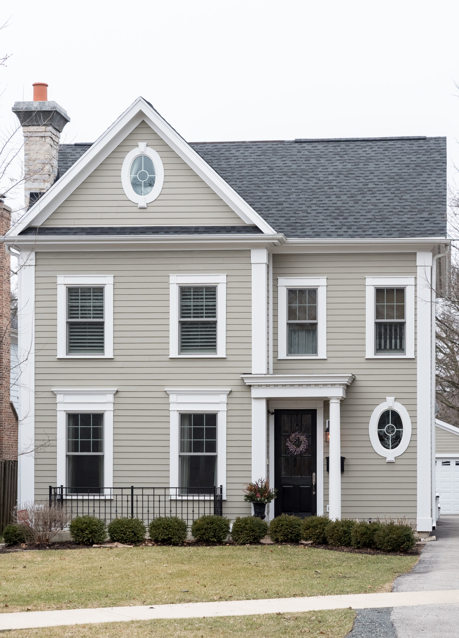 A two-story house with tan siding, crisp white trim, a dark shingled roof, and distinctive oval accent windows